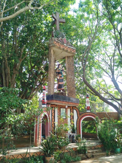 The image depicts a religious shrine or monument situated within a verdant, tree-filled area in An Phu Ward, Vietnam. The structure is tiered, featuring a statue of a woman holding a child under a cross-topped canopy at its highest point. The lower levels incorporate arched gateways, decorated with a patterned motif. Plants and foliage surround the monument, integrated with the landscape. The setting is bathed in sunlight, highlighting the greenery.