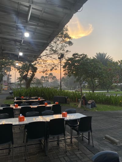 This is an outdoor dining area with multiple tables and chairs set up on a paved surface. The tables are covered with white tablecloths and have orange tissue boxes on them. In the background, there are trees, a building, and a street with cars. The sky is visible with a sunset in progress. The overall scene appears to be a quiet, potentially evening, setting in a semi-urban environment.

Given the provided location of Mekar Bakti, Indonesia, this image likely depicts an outdoor restaurant or cafe in that area. The presence of palm trees in the background suggests a tropical or subtropical climate, consistent with Indonesia. The architecture of the buildings and the general layout of the area are also typical of many Southeast Asian urban or peri-urban environments.