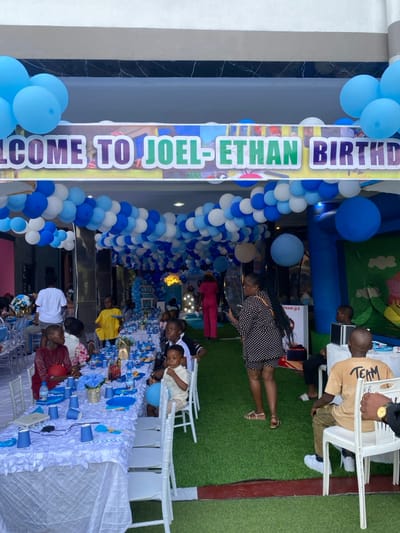 The image shows a birthday party for Joel-Ethan in Bonadikombo, Cameroon (4.048, 9.218). The party is decorated with blue and white balloons and a sign reading "Welcome to Joel-Ethan Birthday". There is a long table with white tablecloth and blue and white decorations. There are people sitting at the table and standing around. One woman is taking a picture with her phone. In the background, there is a bouncy house, some furniture, and an artificial grass area.