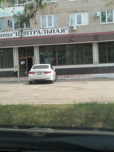 A white sedan is parked on a gravel lot in front of a brick building with large windows. The building has a sign that reads "ЦЕНТРАЛЬНАЯ" which translates to "Central". The license plate on the car is partially visible and appears to be in Russian format. The car is facing away from the building. The weather appears to be overcast. The overall scene depicts an outdoor commercial or administrative area.