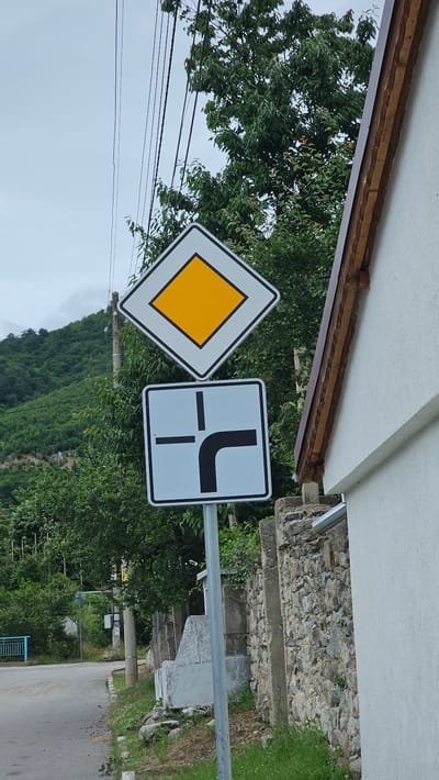 A diamond-shaped sign with a yellow interior and a white border, indicating a priority road, is mounted on a pole. Below it, a square sign with a black border and a light blue interior depicts a T-junction with a right turn from the side road. The signs are positioned next to a stone wall and a white building. The environment is a rural setting with trees and a cloudy sky.