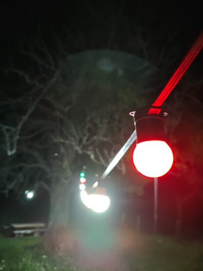 This is a nighttime outdoor photograph, possibly taken in a park or wooded area in Ukeshi, Georgia. The foreground is dominated by a brightly lit red light bulb, part of a string of lights. Behind it, a series of other lights are visible, some appearing green and white, possibly indicating a traffic signal or decorative lighting. The background is dark and indistinct, with faint outlines of trees and branches, suggesting a natural setting. In the middle ground, a bench or picnic table can be seen on the left side of the image, partially illuminated by the lights. The overall impression is one of a dimly lit, natural environment with artificial lighting present.
