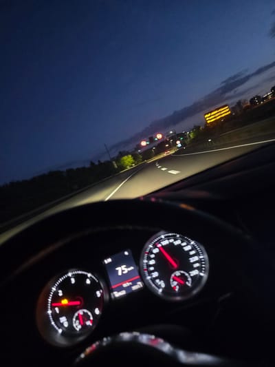 The image is a first-person perspective shot from inside a car driving on a highway at dusk. The dashboard is visible in the foreground, showing a speedometer indicating a speed of 75 km/h. The car's headlights illuminate the road ahead. In the background, the sky is a deep blue, transitioning to lighter hues near the horizon with some clouds. There are streetlights and signs visible along the side of the highway. One prominent digital sign displays text in yellow. The location is identified as Russange, France. The scene depicts a common driving scenario on a highway.