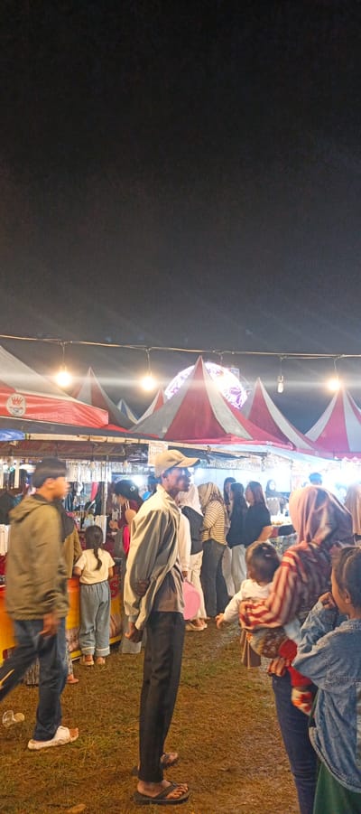This image depicts an outdoor night market scene in Banding Agung, Indonesia. The foreground features several individuals, including a young boy in a green jacket and blue jeans, a girl in a white shirt and light blue pants, and a man in a tan baseball cap and a light-colored shirt with dark pants. Other people are visible in the background, some of whom appear to be shoppers and vendors.
The market is set up under a canopy of red and white striped tents. String lights are hung between the tents, illuminating the scene. In the distance, a large, illuminated spherical decoration is visible, possibly a centerpiece for the market or a decorative element. Stalls are set up beneath the tents, suggesting merchandise is being sold. The ground appears to be covered in grass and is slightly wet, indicating recent rain or damp conditions. The overall atmosphere suggests a lively community event.