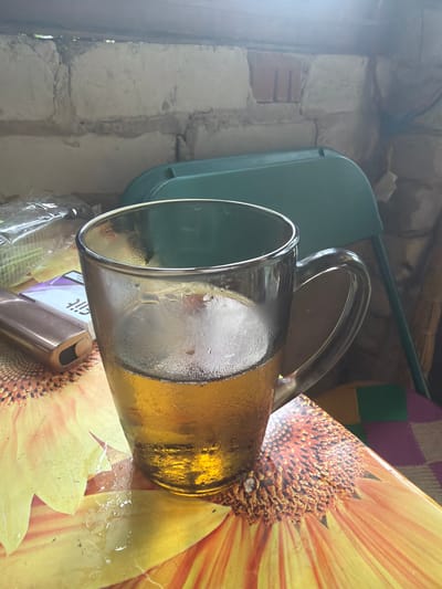 A close-up, eye-level shot shows a glass mug filled with a golden liquid, possibly beer or tea, sitting on a table with a sunflower-patterned tablecloth. The mug is sweating, indicating a cold beverage. To the left of the mug is a metallic, rectangular object that appears to be a cigarette lighter or vape pen, and a pack of cigarettes with purple and white packaging. In the background, a green plastic chair is visible, and the wall behind it is constructed of rough, light-colored bricks, suggesting an outdoor or rustic setting. The overall scene is dimly lit, with the primary light source coming from the right, highlighting the condensation on the glass. There is no indication of the city of Shipovo, Russia, in the image.