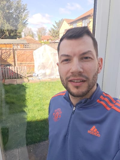An adult male with light skin, short dark hair, and dark stubble is depicted in a medium shot, looking directly at the camera from inside a structure, observing a residential backyard in the Cherwell District, United Kingdom. He is wearing a blue-grey quarter-zip top with orange three-stripe detailing on the shoulders and sleeves. The left chest displays an orange Manchester United crest, while the right chest features an orange Adidas logo. A slight smile is present. The left side of the image shows a reflection of the outdoor scene on a glass surface, likely a window or door. The background features a green grass lawn, a paved area, and is enclosed by a brick wall and a wooden fence. A white, translucent pop-up greenhouse structure is visible towards the rear of the yard. Beyond the property lines, several residential houses with light-colored facades and reddish-brown tiled roofs are visible under a clear blue sky with scattered white clouds, alongside various trees, some with bare branches.