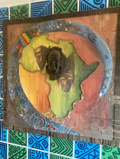 This is a photograph of a piece of art displayed on a wall. The art is a mixed-media collage depicting a stylized map of Africa, with various masks and decorative elements integrated into the composition. The central part of the artwork is a vibrant rendition of the African continent, painted in shades of green, yellow, and orange. Three masks are prominently featured: one at the top center, with intricate dark detailing, and two others placed on the left and right sides of the continent's depiction. The artwork is bordered by a textured frame made of what appears to be ribbed cardboard or similar material, with an outer edge of dark, thin vertical strips. The surrounding wall is tiled with a colorful mosaic of geometric patterns in green, blue, and black. In the lower right corner of the artwork, a signature and date are faintly visible, appearing to read "OLARINSOYE - RIN" followed by "16/12/27" and "2014". The artwork is positioned on a wall in what is identified as Ojere, Nigeria.