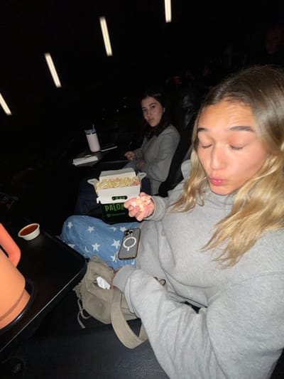 The image captures two females seated in a darkened cinema theater in La Purísima, Mexico. In the foreground, a female with light-colored hair, wearing a grey hooded sweatshirt and blue star-patterned pants, holds a handful of colorful popcorn. A white cardboard popcorn box labeled "PALOMITAS Cinemex" rests on her lap, alongside a smartphone featuring a visible MagSafe ring and a light-colored handbag. To her left, a small white cup containing a reddish liquid is on a black armrest tray. Her eyes are partially closed, and her mouth is slightly puckered. In the background, a second female with dark hair, wearing a light grey jacket, is seated and looking towards the viewer. A white insulated cup with a straw and a dark-colored smartphone are on a side table next to her. The environment features dark cinema seating and multiple linear light fixtures visible against the dark ceiling.