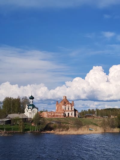 The image depicts a scenic view of Анхимово, Russia, featuring a body of water in the foreground and a clear sky with scattered clouds above. Prominently positioned on the shoreline is a ruined brick building next to a well-maintained white church with a green dome. A small, wooden house is adjacent to the church. A small boat lies near the shoreline on the right side of the image, amidst dried reeds. The environment is a rural landscape with trees dotting the horizon and lush greenery.