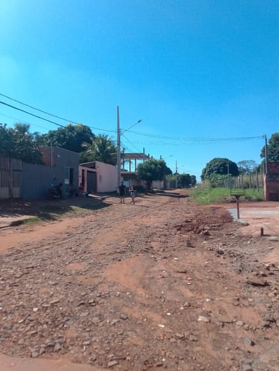 This is a ground-level, eye-level, outdoor, wide shot of a dirt road in a residential area of São Gabriel Do Oeste, Brazil. The sky is clear and blue. The road is unpaved and uneven, with rocks and loose dirt. On the left side of the road, there are houses and fences. One house is painted gray and has a motorcycle parked in front of it. Several people are sitting outside another house, which is painted pink. On the right side of the road, there are more houses and vegetation, including trees and bushes. Two people, a man and a woman, are walking on the right side of the road, away from the camera. There are utility poles and wires running along the road. The scene depicts a typical unpaved residential street in a rural or semi-rural Brazilian town.