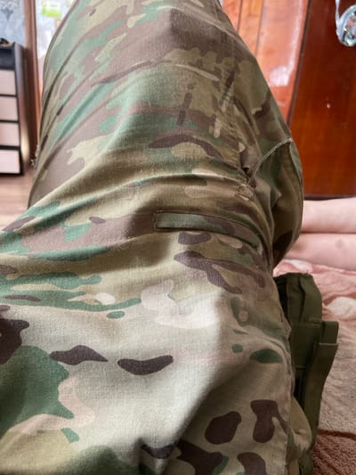 The image shows a close-up view of camouflage pants. The pants are made of a multi-terrain camouflage fabric, featuring patterns of green, brown, tan, and black. The fabric appears to be durable and slightly textured. A pocket or flap is visible on the right side of the pants, made of a darker green material. The pants are creased and folded, suggesting they are being worn or have been recently worn. The background is blurred and out of focus, but it appears to be an indoor setting with wooden furniture and a soft surface like a bed or couch. The lighting is natural and diffused, casting soft shadows on the fabric. The exact location within Valky, Ukraine, cannot be determined from the image alone, as there are no specific landmarks or identifying features present.