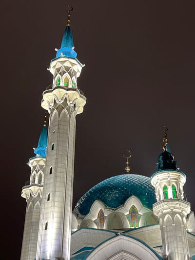 The image displays the Kul Sharif Mosque, located in Kazan, Republic of Tatarstan, Russia, under nighttime conditions. The architectural structure is brightly illuminated against a dark, uniform sky. The foreground features a tall, slender minaret, predominantly white with intricate carvings, a prominent balcony section showcasing green-lit arched windows, and a pointed, blue conical roof crowned with a golden crescent finial. A second, smaller minaret with a similar blue roof and finial is partially visible behind it. To the right, the large main dome of the mosque is visible, characterized by its textured, teal-blue surface and topped by another golden crescent finial. A third, shorter minaret with green-lit windows and a blue conical roof stands on the far right. The mosque's white facade is accentuated by white light, while key windows and dome surfaces exhibit green and blue illumination respectively. No individuals or discernible activities are depicted.