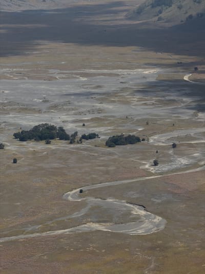This aerial photograph depicts the vast, arid landscape surrounding Cemorolawang, Indonesia, a region known for its volcanic terrain and dramatic natural beauty. The scene is dominated by a winding, dusty riverbed that meanders through a desolate expanse of dry earth, interspersed with patches of scrubby vegetation. The riverbed itself appears to be a mixture of light gray sand and darker, compacted soil, with evidence of past water flow etched into its surface.
Scattered across the landscape are various geological formations, including low-lying mounds and depressions that suggest erosion and the passage of time. In the mid-ground, a dense cluster of dark green trees stands out, forming a striking contrast against the muted tones of the surrounding environment. These trees, likely a resilient species adapted to the harsh conditions, create a small oasis of life in the otherwise barren terrain.
The sky above is partially obscured by shadows cast by unseen elements, possibly clouds or distant mountains. These shadows create a dynamic play of light and dark across the landscape, accentuating the contours and textures of the terrain. The overall impression is one of a remote and rugged natural environment, shaped by the forces of geology and the elements. There are no visible people or man-made structures, reinforcing the sense of untouched wilderness.