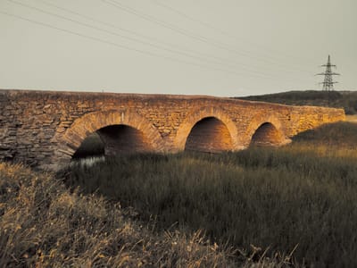 This media file shows an old, arched stone bridge spanning a small body of water. The bridge is made of rough-hewn stones and has four arches. The sun is low in the sky, casting a warm glow on the bridge and the surrounding dry grass. In the background, there is a gently sloping hill covered in dark green vegetation, and a tall, lattice-like electricity pylon with power lines extending across the sky. The scene suggests a rural or semi-rural setting. The light and the dry vegetation indicate it might be late summer or early autumn, likely late afternoon or early evening. There is no visible text in the image.