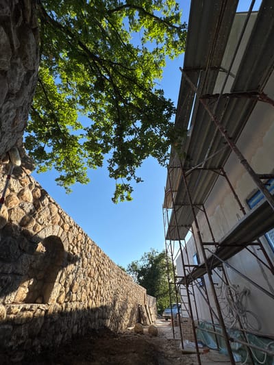 The image shows a construction site on a clear, sunny day, likely in the city of Pelinovo, Montenegro. In the foreground, a rustic stone wall, possibly part of a garden or property boundary, stretches diagonally across the frame. The wall is constructed from rough-hewn, irregular stones of varying sizes and colors, with a subtle arched alcove or feature embedded in it. Sunlight casts strong shadows on the wall, highlighting its texture and the shadows of nearby trees and foliage.
To the right of the stone wall, a building is under construction, with scaffolding erected along its exterior. The building's facade appears to be plastered or painted a light color, with visible construction materials and wiring on its surface. The scaffolding consists of metal poles and horizontal planks, some of which are covered with tarpaulin.
Above, a bright blue sky is visible, with the lush green leaves and branches of a large tree extending into the frame from the upper left. The tree's foliage is dense and vibrant, suggesting it is summertime. In the background, further trees and a silver car are visible, indicating a slightly wider environment beyond the immediate construction area. The ground in the foreground is uneven and appears to be covered with dirt and some construction debris. The overall impression is one of ongoing renovation or building work in a natural setting.