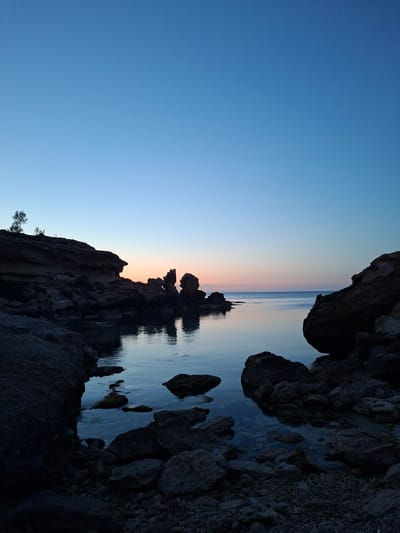 The image presents a serene coastal landscape at either dawn or dusk in Paralimni, Cyprus. The scene captures a tranquil sea bordered by rugged, dark rock formations.
The sky, occupying the upper part of the horizon, displays a soft gradient of colors, transitioning from a deep, clear blue overhead to lighter blue, and then to delicate bands of pink and orange hues right at the horizon. This celestial palette is reflected with remarkable clarity on the calm, still surface of the sea, creating a sense of seamless continuity between the sky and the water.
Along the distant horizon line, several dark, irregular rock formations rise from the tranquil waters, appearing as distinct silhouettes. Their shapes are perfectly mirrored in the glassy sea below, emphasizing the stillness of the water. The foreground and sides of the image are framed by dark, imposing rocky cliffs and outcrops. These rocks exhibit rugged textures and uneven edges, hinting at their ancient formation. On the lower left side, a small, sparse tree or shrub can be seen clinging to the rocky terrain. Pockets of clear water are visible among the rocks in the immediate foreground.
There are no people or specific activities depicted, contributing to the overall sense of peace and natural quiet. The clear weather and smooth water enhance the tranquil and contemplative mood of the scene, highlighting the natural beauty of the Cypriot coastline. There is no visible text.