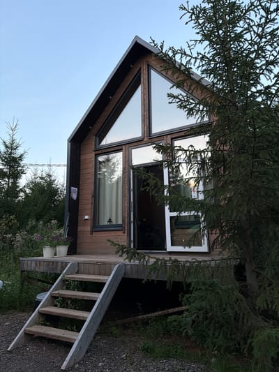 The image shows a wooden cabin with a modern design located in Kuliki, Russia. The cabin features large triangular windows on the upper facade and rectangular windows on the lower level. A wooden deck with a set of stairs leads to the entrance, where a door is ajar. Two white flower pots sit on the deck near the stairs. A large evergreen tree is positioned to the right of the cabin, partially obscuring the view. The surrounding environment is lush with vegetation, including various trees and smaller plants. The sky is a clear, pale blue, suggesting it is either early morning or late evening. There are no people visible in the image. A QR code is affixed to the left side of the cabin's wooden exterior.