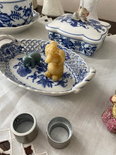 In Osek, Czechia, on a white tablecloth, is a close-up shot of decorative porcelain items and small figurines. In the foreground, two silver cylindrical objects rest on the fabric. Behind them, a white porcelain tray with a blue floral pattern and a lattice edge holds a small grey elephant figurine and a yellow elephant-shaped candle. Further back, a lidded white porcelain box, also adorned with blue floral designs, is topped with a small, light-colored mouse figurine. To the right of the box, a portion of a small decorative object, possibly a figurine, with a pink glittery base and gold accents is visible. The overall scene suggests a collection of decorative collectibles, likely displayed indoors.