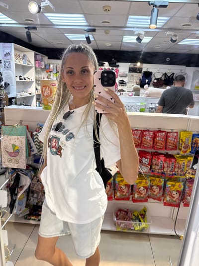 A woman with blonde braided hair smiles at the camera while holding a pink smartphone in a retail store. She is wearing a white t-shirt with a graphic of bears and a can, and light blue denim shorts. Sunglasses are hanging on her t-shirt. The store shelves behind her are stocked with various items, including gift bags, snacks, and clothing. A man in a dark grey t-shirt is visible in the background, facing away from the camera. The environment appears to be a small shop selling a variety of goods. This image was taken in Sosnovtsy, Russia.