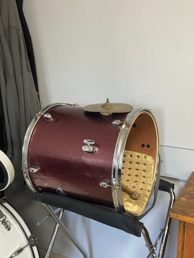 A maroon bass drum is propped on a silver metal stand in front of a white wall. A cymbal rests on top of the drum, and a drumstick lies inside the drum, resting on a yellow, perforated cushion. To the left of the drum, a dark gray jacket hangs on a hanger. A portion of a white drum and a wooden cabinet are visible on the far left and right sides of the image, respectively.