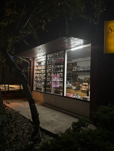 A nighttime exterior shot captures a small shop in Ilidža, Bosnia and Herzegovina, with a brightly lit interior showcasing a wide array of merchandise displayed on shelves and a counter. The shop's large display window is filled with various items, including numerous bags of what appears to be tea or coffee, jars, and decorative boxes. In the foreground, a large tree trunk with sparse leaves leans across the left side of the frame, partially obscuring the building. A paved walkway runs in front of the shop. To the right of the shop, a dark brick facade is visible, and a yellow sign with red lettering, partially obscured, is mounted on the wall. The overall lighting suggests the shop is open for business or at least illuminated for security.