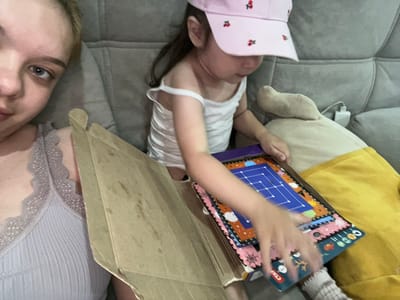 This media file shows a close-up, overhead view of a group of people on a couch. The subjects include a child lying on their back, wearing a white tank top and a pink hat adorned with cherries. Their face is partially obscured, but their eyes are visible. To the left of the child, a person wearing a yellow shirt is partially visible, along with a plush toy. To the right of the child, another person is lying on their back, facing the camera. They are wearing a light blue ribbed top with lace trim. A cardboard box is positioned between the child and the person on the right. The child is holding a colorful board game, possibly a puzzle, with a blue grid pattern. The image appears to be taken indoors, likely in a home setting, given the couch and the presence of a plush toy. The lighting suggests it could be daytime. The overall mood seems relaxed and casual, possibly during playtime or a quiet moment at home in Gorodets, Russia. The board game has some visible text, including "CTA" and numbers.