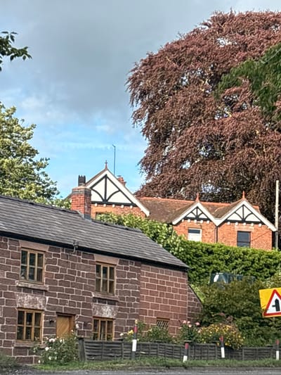 The image depicts a scene in Brown Knowl, United Kingdom, featuring a row of brick buildings and lush greenery under a partly cloudy sky. In the foreground, a dark-tiled roof covers a building constructed of reddish-brown brick with visible mortar lines. This building has multiple windows with wooden frames. To the right, a dark wooden fence borders a grassy area with flowering bushes. Further back, a taller brick building with a pitched roof and decorative white gables is visible, partially obscured by dense green foliage. A prominent, large tree with reddish-brown leaves dominates the upper right portion of the image. A yellow road sign with a black symbol indicating a junction or lane split is positioned on the right side. The environment appears to be a rural or semi-rural setting. No people are present in the image.