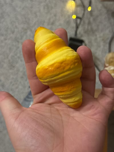 An over-the-shoulder, close-up shot shows a person's left hand holding a miniature croissant-shaped toy. The toy is a bright yellow with orange swirls mimicking the flaky layers of a real croissant. The background is out of focus, but appears to be an indoor setting with a textured gray surface, possibly carpet, and out-of-focus string lights. There are no visible landmarks or indicators of a specific location within Molodezhnyi, Russia.