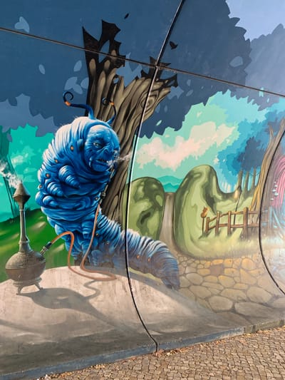 This media file depicts a mural painted on a curved surface, likely a wall or a structure. The artwork features a large, blue, anthropomorphic caterpillar with distinct facial features, including wide eyes and a somewhat smug expression. The caterpillar is shown smoking from a hooka pipe, with smoke emanating from it. An orange tube connects the hooka to a nozzle near the caterpillar's mouth. The caterpillar is depicted against a surreal landscape with large, rounded green shapes that could represent oversized foliage or strange rock formations. A wooden ladder leans against one of these green shapes. The background transitions from a dark blue and black area, suggestive of a night sky or shadowy trees, to lighter blue and green hues, hinting at a dreamlike or fantastical environment. The artwork is situated outdoors, as evidenced by the stone-paved ground visible on the right side, some fallen leaves, and the natural lighting. The time of day appears to be daytime, likely sunny, given the shadows cast. The style of the mural is illustrative and detailed, with a vibrant color palette. The location context of Odivelas, Portugal, suggests this is a public art installation. There is no visible text within the media file.