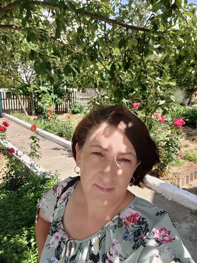 A medium shot, eye-level, outdoor selfie shows a woman in Kryukov, Russia, on a sunny day. She is facing the camera and smiling slightly. She has brown hair, fair skin, and is wearing a light green shirt with a floral pattern. She has hoop earrings in her ears. The background shows a garden with trees, flowers, and a wooden fence. The woman is standing on a paved path. Sunlight casts shadows on her face and the surrounding garden.