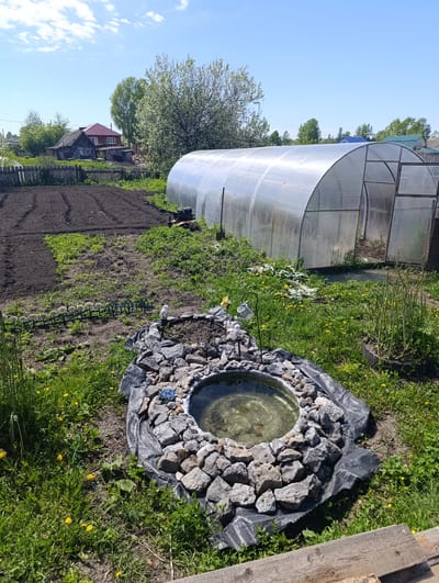 This media file captures a bright, sunny rural garden or backyard scene, likely in Yaya, Russia. The landscape features a mix of cultivated areas and natural growth, bathed in what appears to be mid-morning or early afternoon sunlight under a clear blue sky with scattered white clouds.

In the prominent right-hand side of the image, a large, arched greenhouse with translucent polycarbonate panels and a metal frame stands, its door open. To the left of the greenhouse, a substantial section of dark, tilled soil is visible, meticulously prepared in rows, suggesting active gardening and planting. A small rototiller or garden tiller is parked between the tilled soil and the greenhouse.

In the foreground, a circular, in-ground pond or water feature serves as a decorative focal point. It is surrounded by an arrangement of grey rocks and dark geotextile fabric, and contains clear water. Small garden ornaments, including a white ceramic bird and a miniature metal watering can on a stand, adorn the pond's edge, along with a blue decorative flower among the rocks. Further left, a line of white floral decorations is placed along a green border. The ground around these features is covered in green grass dotted with yellow dandelions.

Beyond the garden plot, a rustic wooden picket fence marks a boundary. In the distance, traditional wooden houses with reddish and blue roofs are visible, nestled amidst various trees, some of which appear to be in bloom with white blossoms. No individuals are present in the scene, which conveys a sense of peaceful rural activity.