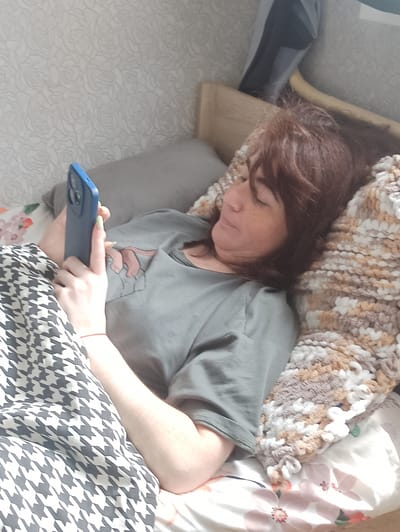 A woman with brown hair and freckles is lying in bed, holding a blue smartphone in her right hand. She is wearing a grey t-shirt with a graphic design of hands on the pocket. A blanket with a houndstooth pattern is draped over her left side, and a chunky knit blanket with brown, white, and grey yarn is on her right. The background shows a wall with a subtle rose pattern wallpaper. The scene is indoors, likely a bedroom. The location in the city of Verkhnii Lars, Russia, is unknown.