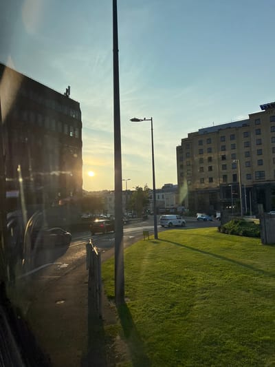 The image captures a street scene in the city of Derry/Londonderry, United Kingdom, during what appears to be sunset. A large, dark building, possibly an office block, dominates the left side of the frame, with the setting sun reflecting off its windows. Across the street, a light-colored, multi-story building, likely a hotel, stands prominently. The street is moderately busy with cars, including a white van, a dark car, and a blue car visible. A well-maintained grassy area is situated in the foreground on the right, with a tall lamppost casting a shadow. The sky is clear with a soft gradient of blue and yellow hues indicating the setting sun. The overall impression is of an urban environment bathed in the warm light of dusk.