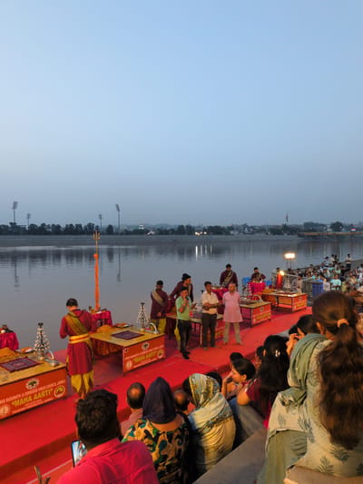 The image depicts a Ganga Aarti ceremony taking place on the banks of the Tawi River in Jammu, India, possibly at the Mubarak Mandi complex. In the foreground, a group of people are seated on red carpeting, with some individuals standing and participating in or observing the ceremony. One man in a pink tunic and another in a red and yellow robe are prominent. Several tables with religious items and offerings are set up. In the background, the Tawi River stretches out, with a cityscape visible on the opposite bank, including lights and a bridge. Tall floodlights and trees line the distant riverbank, and the sky is a pale blue, suggesting either dawn or dusk. The ceremony appears to be well-attended, with a crowd gathered to witness the event.