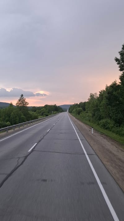 The image shows a straight, paved road leading into the distance. The road has white lane markings and a metal guardrail on the left side. The right side of the road is bordered by a dirt shoulder and lush green trees. In the background, rolling hills covered in trees are visible under a hazy sky with hints of pink and orange, suggesting a sunset or sunrise. The overall environment is rural and natural. There are no people or specific actions depicted. The location is not Uril, Russia, as there is no information to support this claim.