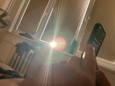 In Acones Dārzs, Latvia, an indoor scene unfolds with strong sunlight streaming through a window, creating significant lens flare and glare. A person's hand is visible in the foreground, holding a dark-colored smartphone. The phone's screen displays text. Behind the hand, a window with blinds is prominent, with the sun's rays emanating from it. A dark green thermos is positioned on the windowsill. To the left of the window, a radiator with some folded clothes draped over it is visible. Further left, a fan with a metal grill is mounted on the wall. The overall environment suggests a domestic setting within the specified location.