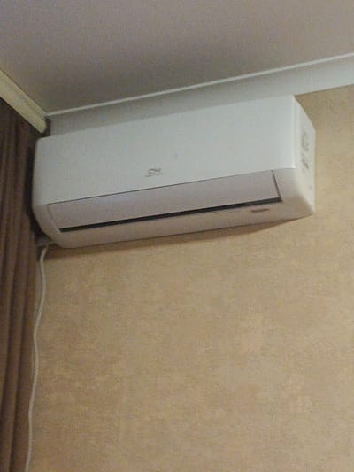 A white air conditioner unit is mounted on a beige, textured wall near the ceiling. A white electrical cord hangs from the unit, descending along the wall. To the left of the air conditioner, a section of a dark brown curtain is visible. The ceiling is a light color, and a white trim runs along the junction of the wall and ceiling. The image appears to be taken indoors, likely in a residential or commercial space. The location within Balakliya, Ukraine is not discernible from the visual information in the image.