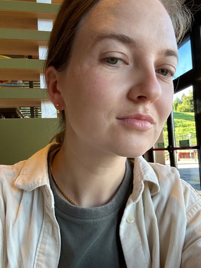 A close-up, head-and-shoulders photograph depicts a young adult female in an indoor commercial setting, located in Lipomo, Italy. The female has fair skin, light-colored eyes, and a neutral facial expression, looking towards the camera. She wears a dark olive-colored crew-neck shirt beneath an unbuttoned light beige corduroy shirt, a thin gold chain necklace, and a small gold hoop earring with square indentations on her left ear. The indoor background to her left is blurred with horizontal wooden or light-colored slats. To her right, a large window reveals a sunny outdoor scene featuring a green hillside with trees, a red metal barrier, and a green rectangular sign with white text, likely an exit indicator.