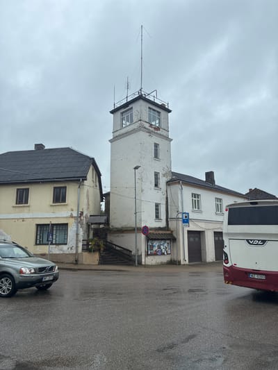 The image depicts a street scene in Sabile, Latvia, under an overcast sky. In the foreground, the wet asphalt suggests recent rain. A silver Volvo SUV is parked on the left, facing away from the camera. To the right, the rear of a white and red VDL bus is visible, also facing away.

The central focus of the image is a tall, white, multi-story building that appears to be an old fire watchtower. It has several windows and an antenna at the very top. Adjacent to the tower is a pale yellow, two-story building with a dark gray roof. A set of outdoor stairs leads up the side of the yellow building. There's a bulletin board on the side of the tower building, displaying various notices. Blue and white road signs are also visible, one indicating a parking area and another a pedestrian crossing. Behind the main buildings, another structure with a dark roof and chimney can be seen. The overall impression is one of a quiet, somewhat weathered town center.