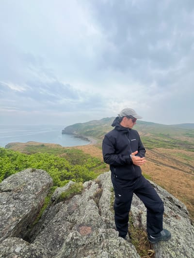 The image depicts a person standing on a rocky cliff overlooking a body of water. The individual is wearing a dark-colored jacket and pants, a cap, and dark shoes. They are positioned at an angle, appearing to lean towards the edge of the cliff. Behind them, the landscape consists of grassy fields and patches of green vegetation. The sky is overcast with a layer of clouds. The body of water extends into the distance, with a visible coastline and small bay. Based on the context of the city, the location is likely a coastal area in Gvozdevo, Russia.