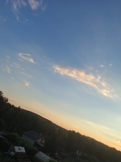 This is a photograph of a sloped residential area in Dobev, Czechia, taken during the day. The image features a clear blue sky with scattered wispy clouds and a sunset gradient of pale orange and yellow near the horizon. The foreground is dominated by a dense forest that covers a hillside, with a few houses and some gardening structures visible amidst the trees. A prominent house with a dark roof and a brick chimney is situated on the slope. In the lower left corner, there are two greenhouses, one with white flowers inside. The overall impression is of a rural or semi-rural setting with residential structures integrated into the natural landscape.
