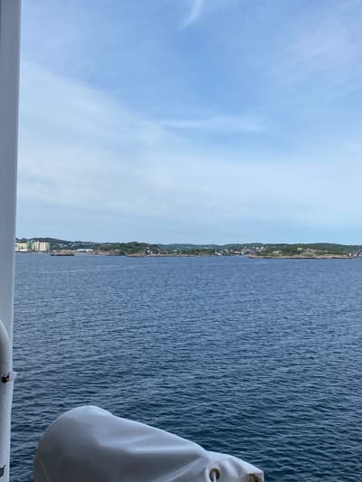 The image captures a view of Stavern, Norway, from a boat or ferry. The scene is divided into two distinct halves. On the left, a pale blue sky with faint wispy clouds dominates the frame, suggesting either early morning or late afternoon. A white railing runs horizontally across the bottom of this portion, indicating the vantage point is from a vessel.
The right half of the image showcases the deep blue water of a body of water, likely the sea. The water is textured with small ripples, indicating a gentle breeze or movement. A white object, possibly a boat cover or part of the vessel's superstructure, is visible in the lower right corner.
Along the horizon, a green, hilly coastline is visible, dotted with a scattering of white buildings that make up a town or village. The presence of trees and vegetation suggests a temperate climate. The overall impression is one of a serene and picturesque coastal landscape. No visible text is present in the image. The lighting suggests it is daytime, with the sun likely low in the sky, creating a soft, diffused light.