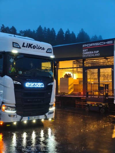 The image depicts a nighttime or dusk scene in Flatschach, Austria, on a wet, reflective paved surface. A white Scania semi-truck, featuring illuminated headlights, fog lights, and a blue-white 'SCANIA' grille logo, is positioned in the foreground. The truck's sun visor is marked 'LiKoLine', and its front bumper displays German license plate 'BCH SN 403'. To the right, a dark, temporary building or tent structure with large glass panels is brightly lit from within by warm yellow light. The structure's facade is branded with 'PORSCHE' in red and white, and 'SIXT CARRERA CUP DEUTSCHLAND' in white. Inside, multiple light-colored wooden crates are stacked. A yellow and black scissor lift is parked on a raised platform adjacent to the building. A dark forest line is visible in the background against a dark blue sky. No individuals are present.