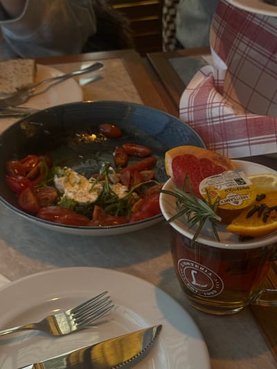 The scene depicts an indoor dining table at a "L'Osteria" restaurant in Mysiadło, Poland. In the midground, a dark blue bowl contains a partially consumed salad consisting of halved cherry tomatoes, white cheese (likely mozzarella or similar), and green arugula leaves. To the right, a clear glass mug branded "L'Osteria" holds a light brown liquid, presumed to be tea or herbal infusion. A small white saucer rests atop the mug, holding garnishes: a slice of pink grapefruit, a small plastic container of "IMKERGOLD BRETSAMER HONIG" (honey), a lemon slice, an orange slice studded with cloves, and a sprig of rosemary. In the foreground, a white ceramic plate is set with a silver fork on the left and a silver serrated knife on the right. The tabletop is a light, neutral color, possibly stone or light wood grain. In the background, a red and white checkered napkin is visible on the right, and blurred figures of individuals seated at other tables are discernible, along with additional cutlery and a white plate, indicating multiple diners in the restaurant environment.