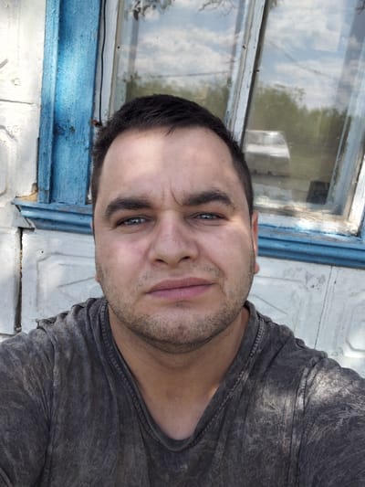 This is a selfie taken by a young man in Ketrysanivka, Ukraine. The man is wearing a dark grey t-shirt. He is positioned in the center of the frame, with his face tilted towards the camera. His expression is a grimace or a scowl, with his eyebrows furrowed and his mouth slightly open. His eyes are a striking blue and are looking directly at the camera. He has short, dark hair and a light stubble on his face. The background of the image shows a weathered, light-colored wall and a rustic wooden window frame painted in a peeling blue. The window glass reflects the sky and some greenery, suggesting it is daytime and likely outdoors. The overall impression is one of candidness, possibly capturing a moment of mild discomfort or exertion. There is no visible text in the image.