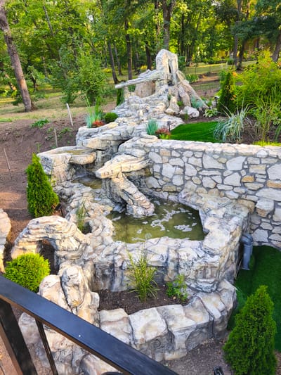 The image shows an artificial waterfall and pond feature surrounded by landscaping, likely in a park or garden setting. The waterfall is constructed from light-colored, textured rocks, mimicking natural stone. Water cascades down several levels into a pool at the bottom, which appears slightly murky and has some bubbling or agitation on the surface. The pond is contained by a wall of similarly constructed rocks and a section of rough-hewn stone wall. Several small evergreen shrubs and other greenery are planted around the feature, and artificial turf is visible in some areas. In the background, mature trees with lush green foliage suggest a wooded environment. The image is taken from a slightly elevated perspective, looking down at the waterfall feature, with a dark metal railing in the foreground. The location is identified as being in the city of Stroiesti, Moldova.