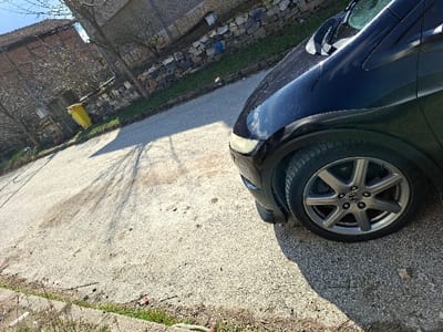 The image depicts an outdoor scene on a clear, sunny day in Rakitovo, Bulgaria, likely a residential street. The primary subject is the front right quarter of a black vehicle, specifically a Honda Civic, visible from the headlight and front fender to the wheel and tire, occupying the right side of the frame. The vehicle features multi-spoke grey alloy wheels with a visible Honda logo on the hubcap. The paintwork appears clean but shows some minor dust or reflections.

The vehicle is parked or positioned on a light-colored, granular paved surface, likely concrete or asphalt, which covers the majority of the ground plane. This surface exhibits some dirt, scuff marks, and scattered debris. Strong, elongated shadows from unseen objects or bare trees are cast across the pavement, extending from the upper right towards the lower left.

In the mid-ground and background, the scene transitions to a residential setting. A concrete curb with sparse green and dry vegetation is visible along the lower left edge. Further back, a bright yellow refuse bin stands near a rough stone wall, which forms a boundary. Behind the wall, a house with a reddish-brown tiled roof and light-colored walls is partially visible, obscured by several bare deciduous trees. The sky is bright blue and cloudless. No individuals are present within the frame.