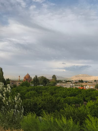 This media file depicts a rural landscape in Artashat, Armenia, under a cloudy sky. The scene is dominated by lush green foliage in the foreground, possibly an orchard or dense vegetation. Behind this, a cluster of buildings is visible, including a distinct, domed red-brick church with a tall, slender tower beside it, suggesting a religious or historical site. Further in the background, more buildings and structures are scattered across the landscape, interspersed with trees. In the distance, rolling hills and mountains form the horizon, with one prominent, lighter-colored hill. The sky is filled with dramatic, layered clouds, indicating overcast or perhaps approaching weather. The lighting suggests it is either early morning or late afternoon. The overall impression is one of a peaceful, somewhat remote, and verdant setting. There is no visible text in the image.