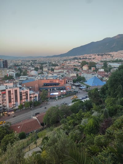 This is an eye-level, horizontal, medium shot of a cityscape with mountains in the background. The photo was taken at dusk, as indicated by the soft, warm light and the gradient of the sky, which transitions from a pale blue to a warm yellow near the horizon. The weather appears clear and calm.

The scene depicts a dense urban area with a mix of modern and older buildings. In the foreground, lush green trees and foliage suggest a park or elevated viewpoint overlooking the city. A paved path with a black metal railing is visible, winding down into the greenery.

Mid-ground, the city unfolds with buildings of various sizes and architectural styles. A prominent feature is a large, pyramid-shaped structure with a bright blue glass exterior. Nearby, a modern shopping center or complex with large windows and signage is visible. There are several yellow taxis on the road, and a few people can be seen walking on the sidewalks. A Turkish flag is displayed on the side of a building. The road is busy with some vehicle traffic.

In the background, the city extends towards rolling hills and then to a large mountain range, which is partially covered in vegetation and appears to be bathed in the warm evening light. The overall mood is one of peaceful transition from day to night in a vibrant urban setting nestled against a natural landscape.

Visible text includes "Zafer Plaza" and "iStanBUL Kurs".