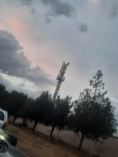 The image depicts a cell tower situated in the city of Hulbuk, Tajikistan. The tower, a tall metal structure with multiple antennas, is positioned to the left of center in the frame, angled slightly against a cloudy sky. In the foreground, a row of dark green trees lines a pathway, with a light-colored wall visible behind them. A portion of a white car is visible on the far left of the image, with its headlights illuminated. The ground is covered in dry grass and dirt, suggesting a somewhat arid environment. The sky is overcast with a mix of gray and pink clouds, indicating either a sunrise or sunset. There are no people or animals visible in the scene.